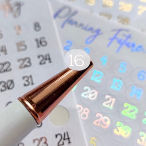 May include: A white pen with a rose gold tip is positioned near calendar stickers. The stickers feature numbers and the text "Planning Future etsy" in a holographic design. The number 16 is highlighted on a sticker.