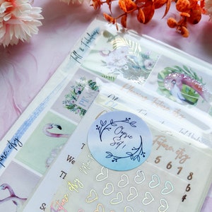 Puede incluir: Una colección de pegatinas y accesorios para planificadores. La imagen presenta pegatinas con diseños de flamencos y flores, junto con pegatinas en forma de corazón y números. El texto "Planning Future.etsy" y "Oopsie Set!" es visible.