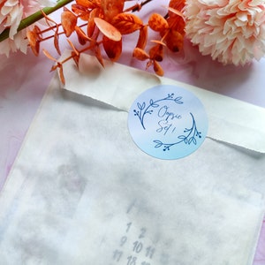 以下が含まれることがあります： 青い花の模様で「Oopsie Set！」と書かれた丸いステッカーで封印された白い紙の封筒。封筒は部分的に透明で、数字の書かれたカレンダーが見えます。背景には、オレンジとピンクの装飾的な花があります。