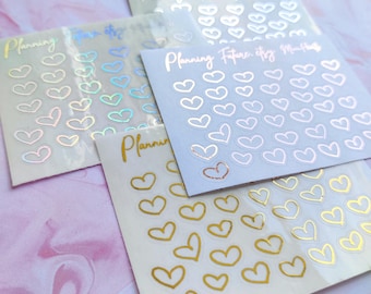 May include: Four sheets of heart-shaped stickers in various colors, including gold, iridescent, and rose gold. Each sheet has the words "Planning Future etsy Months" printed on it. The stickers are arranged on a pink and white marbled surface.