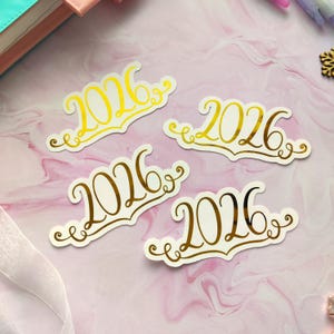 May include: Four stickers with the number 2026 in a decorative font. Two stickers are gold on white, and two are brown on white. The stickers have a decorative border and are arranged on a pink and white marble surface.