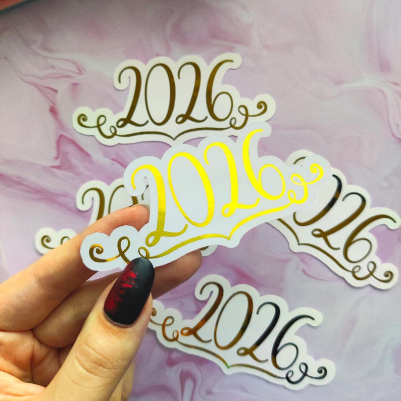 May include: White stickers with the gold text "2026" in a decorative font. The stickers are held by a hand with a black and red nail design. Several stickers are visible against a pink and white background.