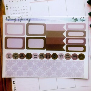 May include: A sticker sheet featuring various planner labels in shades of brown, mauve, and cream. The sheet includes rectangular boxes, flag-shaped markers, rounded icons with black line drawings, and a wide strip with a repeating scalloped pattern.