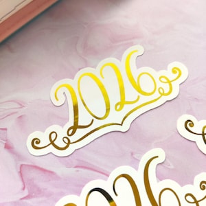 May include: Gold foil sticker with the year 2026 in a decorative script font. The sticker is on a white background with a scalloped edge. The background is a pink and white marbled pattern.