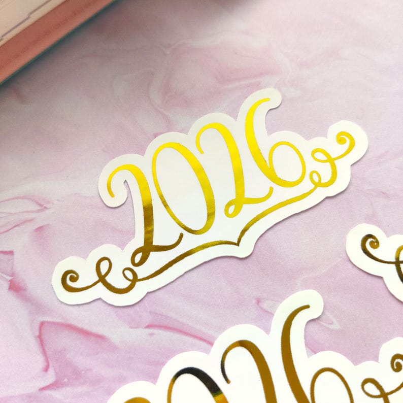 May include: Gold foil sticker with the year 2026 in a decorative script font. The sticker is on a white background with a scalloped edge. The background is a pink and white marbled pattern.