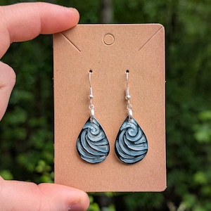 May include: A pair of teardrop-shaped earrings with a swirling blue and white design. The earrings hang from silver-colored hooks and are displayed on a brown card. The background is a blurred view of green foliage.