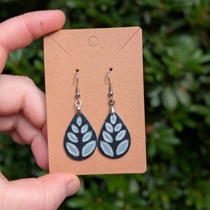 May include: A pair of teardrop-shaped dangle earrings featuring a black base with a speckled texture and a light blue leaf-like pattern. The earrings are attached to a rectangular brown cardboard display card with silver-toned metal hooks.