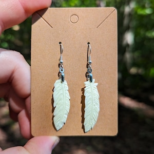 Puede incluir: Un par de pendientes de plumas plateadas de color verde claro. Los pendientes se muestran en una tarjeta marrón.