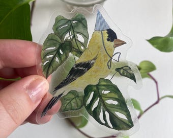 May include: A clear sticker featuring a yellow bird with a black head and a blue party hat, perched on green leaves. The bird has a white bow tie. The sticker is transparent and has a watercolor style.