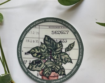 May include: A round sticker with a green border and the text "Support your local library" repeated around the edge. The sticker features a drawing of a potted plant with large, green leaves. The text "Borrower's Name" and a number are visible on the sticker.