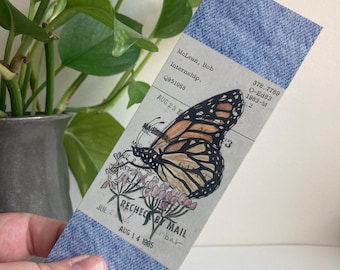 May include: A denim bookmark featuring a monarch butterfly illustration with pink flowers. The bookmark includes text: "McLean, Bob Internship. Q951068 AUG 23 1985". The bookmark is held in a hand.