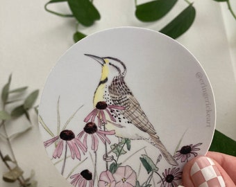 May include: A round sticker featuring a watercolor illustration of a yellow-breasted chat perched on a branch with pink and purple wildflowers. The sticker has a white background and the artist's Instagram handle '@carringrickart' is printed in the bottom right corner.