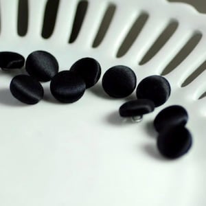 May include: A collection of black, round, fabric-covered buttons. The buttons are scattered on a white, textured surface. The buttons have a shiny, satin-like appearance and are likely for sewing or crafting projects.