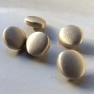 May include: Five small, round, light-coloured buttons are scattered across a white surface. The buttons have a slightly raised, rounded profile and a matte finish, suggesting a natural material like wood or bone. The lighting creates subtle shadows.
