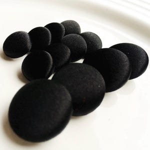 May include: A collection of black, round, fabric-covered buttons. The buttons are arranged in a cluster, showcasing their smooth, matte texture. These buttons are ideal for sewing projects, adding a classic touch to garments and crafts.
