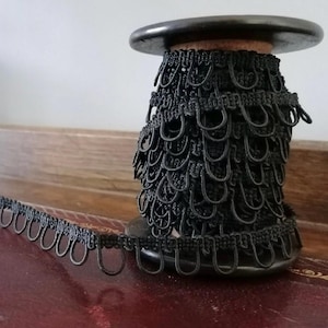 Op de afbeelding: Een spoel met zwarte lusvormige sierrand. De sierrand bestaat uit een reeks kleine, verbonden lussen, die een decoratieve rand vormen. De spoel is donkerbruin met een houten kern. De sierrand is uitgerold op een donkere ondergrond.