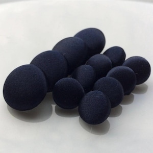 May include: A selection of navy blue fabric-covered buttons in different sizes, grouped together. The buttons have a soft, textured finish and are set against a white backdrop. The buttons are circular and have a slightly raised profile.