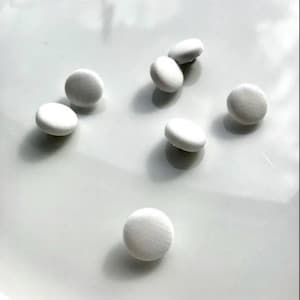 May include: Seven white, fabric-covered buttons are scattered across a white surface. The buttons are circular and have a slightly raised profile. Shadows indicate a light source from above, highlighting their texture.