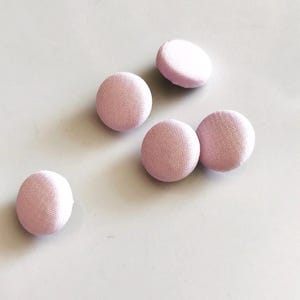 May include: Five light pink fabric-covered buttons. The buttons are round and have a textured surface. The buttons are scattered on a light gray surface.