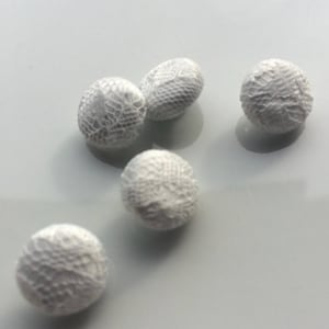 Puede incluir: Cinco botones blancos y redondos con una delicada textura similar al encaje. Los botones están dispersos sobre una superficie clara, mostrando su intrincado diseño, ideal para proyectos de artesanía o decoración de prendas.
