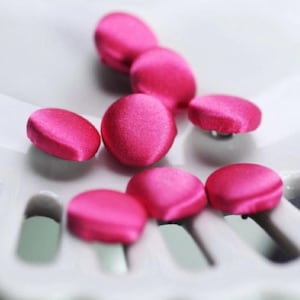 May include: Close-up of several bright pink, satin-covered buttons. The buttons are round and slightly domed, with a smooth, shiny texture. They are arranged on a white surface, suggesting a craft or sewing project.
