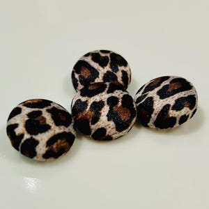 May include: Four round buttons with a leopard print design. The buttons feature a tan base with dark brown and black spots. The buttons are made of a soft, textured fabric.