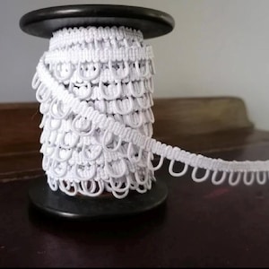 Puede incluir: Un carrete de flecos blancos con un diseño de bucles. El ribete está enrollado firmemente alrededor de un carrete negro, con una parte del ribete que se extiende hacia afuera. El ribete está hecho de un material suave y flexible.