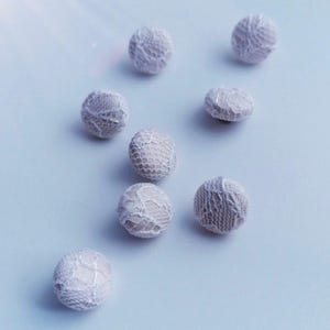 Könnte beinhalten: Neun kleine, runde, cremefarbene Knöpfe. Jeder Knopf ist mit einem zarten, durchbrochenen Spitzenmuster bedeckt, das eine strukturierte Oberfläche erzeugt. Die Knöpfe sind auf einem hellblauen Hintergrund verstreut, was auf ein Bastel- oder Nähprojekt hindeutet.