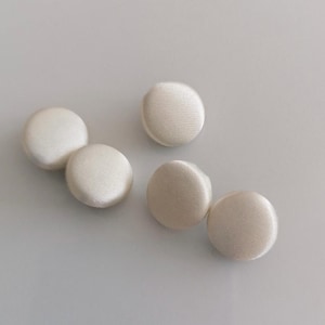 Könnte beinhalten: Fünf elfenbeinfarbene, stoffbezogene Knöpfe. Die Knöpfe sind rund und haben ein leicht erhabenes, gepolstertes Aussehen. Der Stoff hat einen subtilen Glanz, der auf Satin oder Seide hindeutet. Die Knöpfe sind auf einer schlichten, hellen Oberfläche angeordnet.