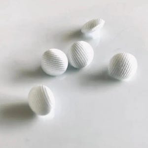 May include: Five white, spherical buttons with a ribbed texture are scattered on a white surface. One button is partially visible, showing its underside. The buttons appear to be made of fabric or a similar material.