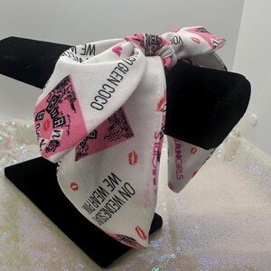 May include: White and pink bandana with black text that reads "We wear pink on Wednesdays" and "You go Glen Coco". The bandana is tied in a knot and has pink lips printed on it.