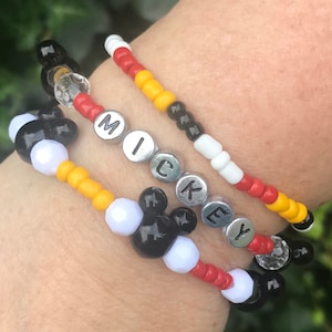 May include: A multi-layered beaded bracelet with black, white, red, and yellow beads. The bracelet features the word "MICKEY" spelled out in silver beads and a Mickey Mouse head design.