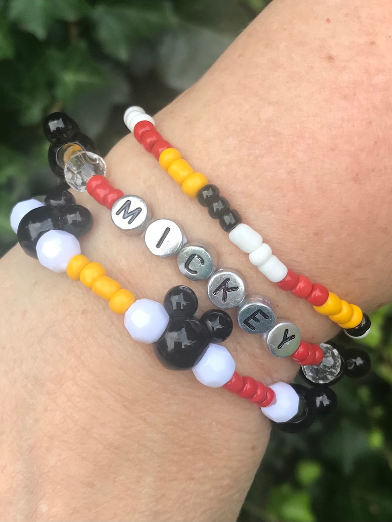 May include: A multi-layered beaded bracelet with black, white, red, and yellow beads. The bracelet features the word "MICKEY" spelled out in silver beads and a Mickey Mouse head design.