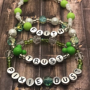 Könnte beinhalten: Drei übereinanderliegende Armbänder mit grünen und weißen Perlen. Das obere Armband hat das Wort "FAITH" in silbernen Perlen geschrieben. Das mittlere Armband hat das Wort "TRUST" in silbernen Perlen geschrieben. Das untere Armband hat die Worte "PIXIE DUST" in silbernen Perlen geschrieben.