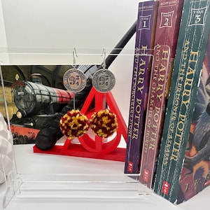 May include: Silver-toned earrings with a "9 3/4" emblem and red and gold pom-poms. The earrings are displayed in front of a red triangular stand and a backdrop of Harry Potter books. A decorative plant and a train image are also visible.
