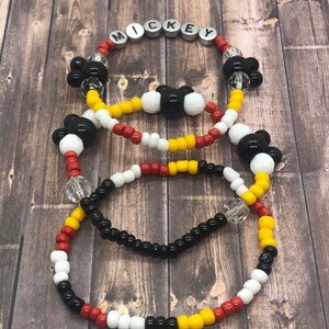 May include: Three beaded bracelets with Mickey Mouse head charms. The top bracelet has the word "MICKEY" spelled out in silver beads. The bracelets are red, yellow, white, and black.