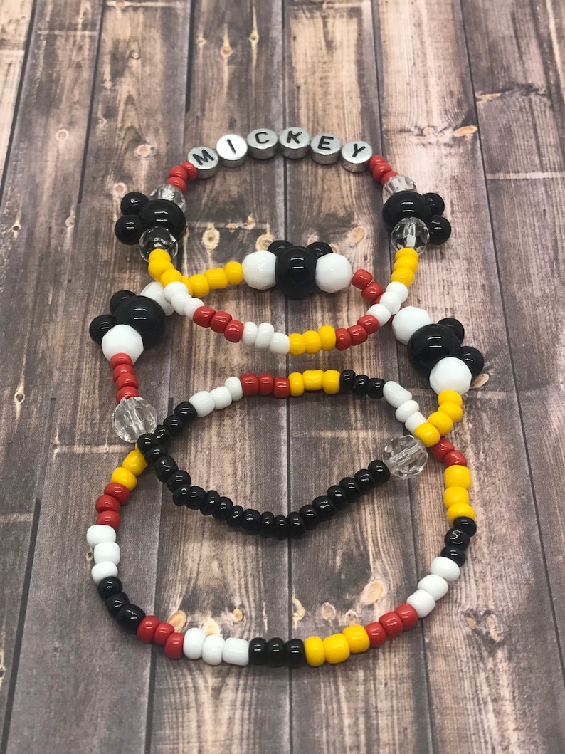 May include: Three beaded bracelets with Mickey Mouse head charms. The top bracelet has the word "MICKEY" spelled out in silver beads. The bracelets are red, yellow, white, and black.