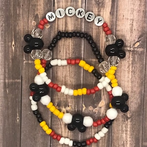 May include: Three beaded bracelets with Mickey Mouse charms. The top bracelet has silver letter beads spelling out "MICKEY". The bracelets are red, white, yellow, and black.