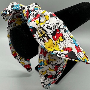 Op de afbeelding: Een witte haarband met een grote strik met een kleurrijk patroon van Mickey Mouse, Minnie Mouse, Donald Duck en Goofy. De haarband wordt gepresenteerd op een zwarte standaard.
