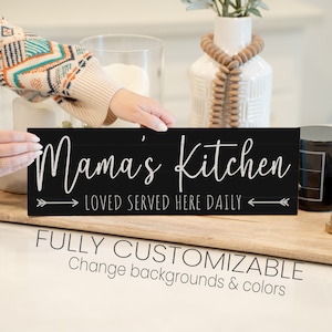 May include: Black wooden sign with white text that reads "Mama's Kitchen Loved Served Here Daily" with arrows on either side. The sign is being held by a hand.