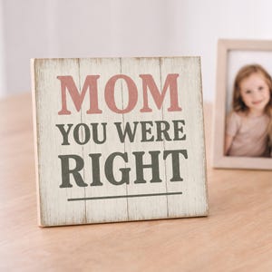 May include: A wooden sign with the words "MOM YOU WERE RIGHT" in pink and gray lettering. The sign has a distressed, rustic look and is placed on a wooden surface. A framed photo is in the background.