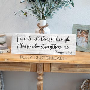 May include: White wooden sign with black text that reads "I can do all things through Christ who strengthens me. -Philippians 4:13". The sign is sitting on a wooden table.