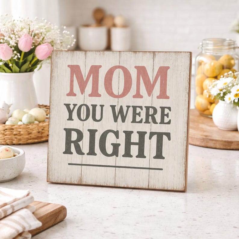 May include: A square wooden sign with the words "MOM YOU WERE RIGHT" in pink and gray lettering. The sign has a distressed, whitewashed finish and is displayed on a countertop with flowers and other decorative items.