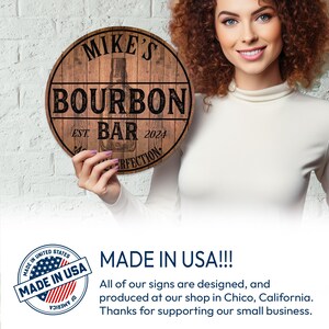 May include: A round wooden sign with the text "MIKE'S BOURBON BAR EST 2024" in black lettering. The sign features a bourbon bottle illustration. The sign is held by a person.