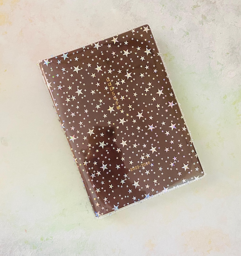 May include: A brown leather-bound journal with a gold foil design of stars and the text "日々を丁寧に" (meaning "Live each day carefully"). The journal is dated 2022-2026.