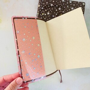May include: Two open notebooks with pink and brown covers. Both covers have a holographic star pattern. The notebooks are open to blank pages.