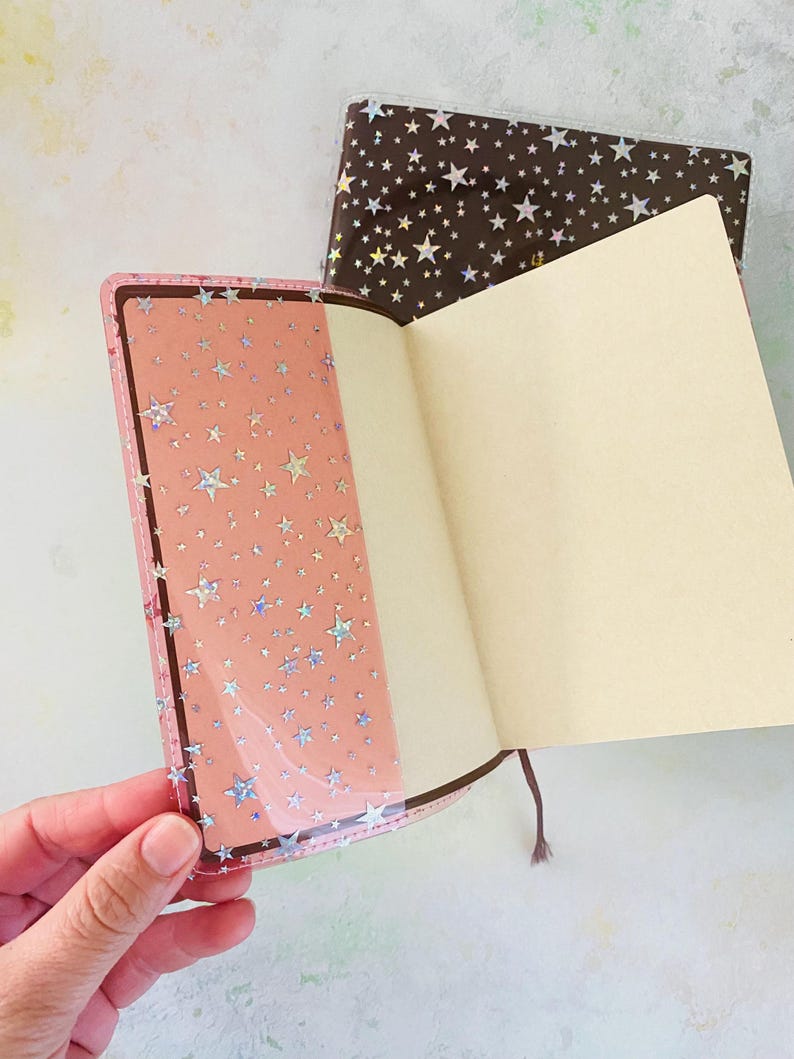 May include: Two open notebooks with pink and brown covers. Both covers have a holographic star pattern. The notebooks are open to blank pages.