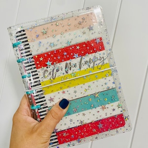 May include: Clear plastic planner cover with a colourful striped design and holographic stars. The cover has the text "Color Me Happy" written on it.