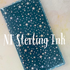 May include: A rectangular, teal-colored notebook cover with a clear overlay. The cover is decorated with a pattern of silver and iridescent stars. The text "NI Sterling Ink" is printed in white across the front.