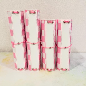 May include: Four journals in varying sizes, featuring a pink and white chequered pattern. The journals are bound with pink elastic and silver eyelets. The spines are white. The overall design is a mix of pink and white.
